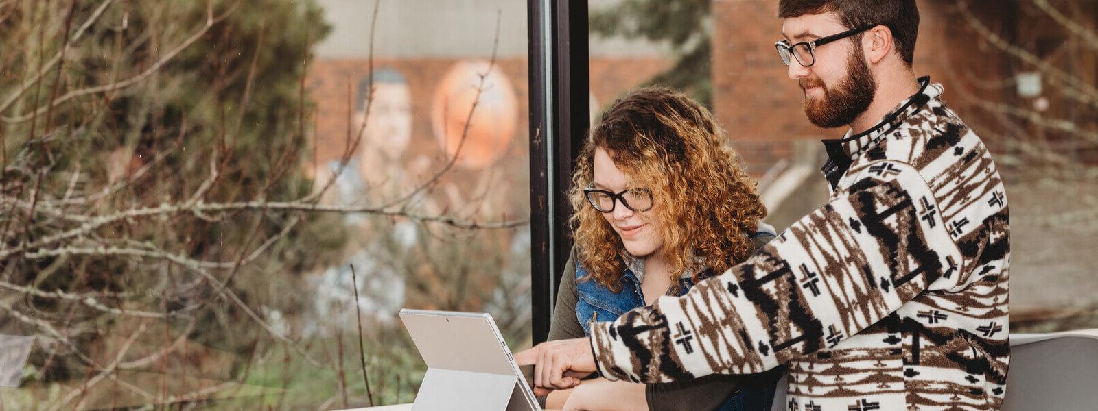 Male student helping and pointing to tablet screen of female colleague